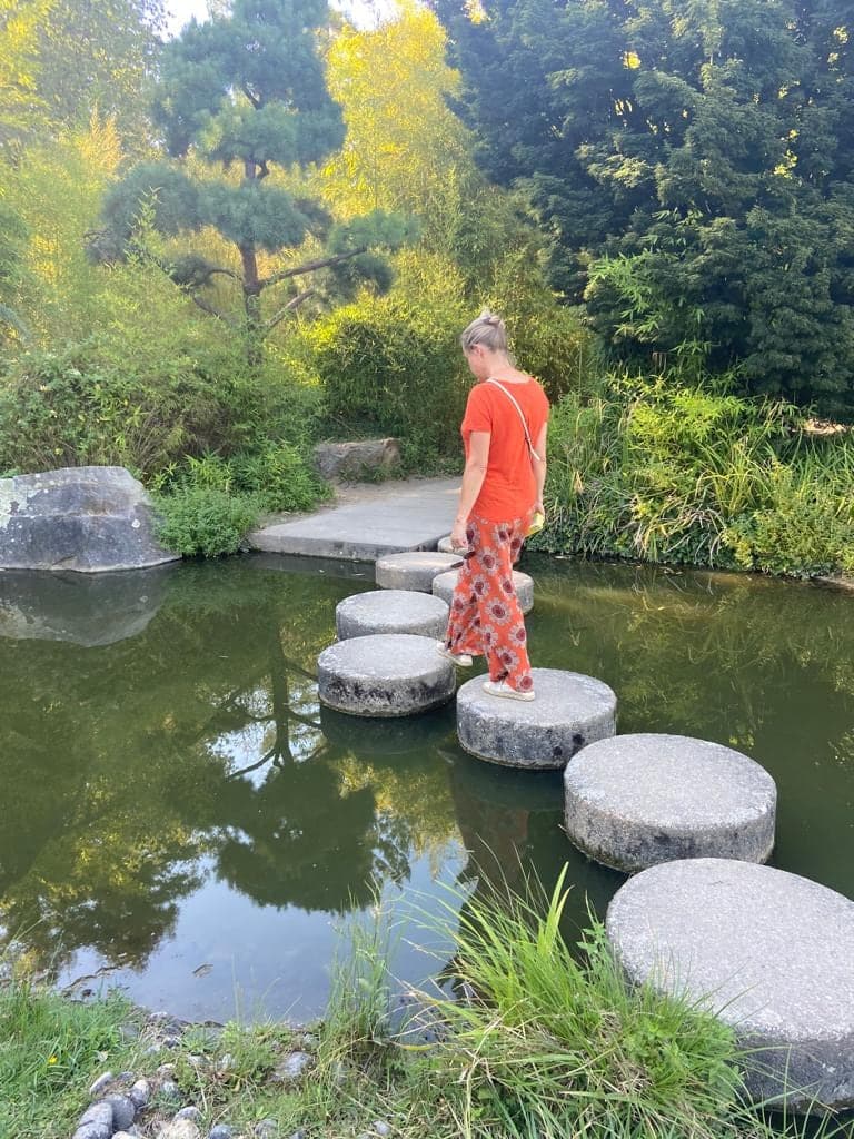 Catherine traverse un étang sur des pas japonais, dans un jardin verdoyant